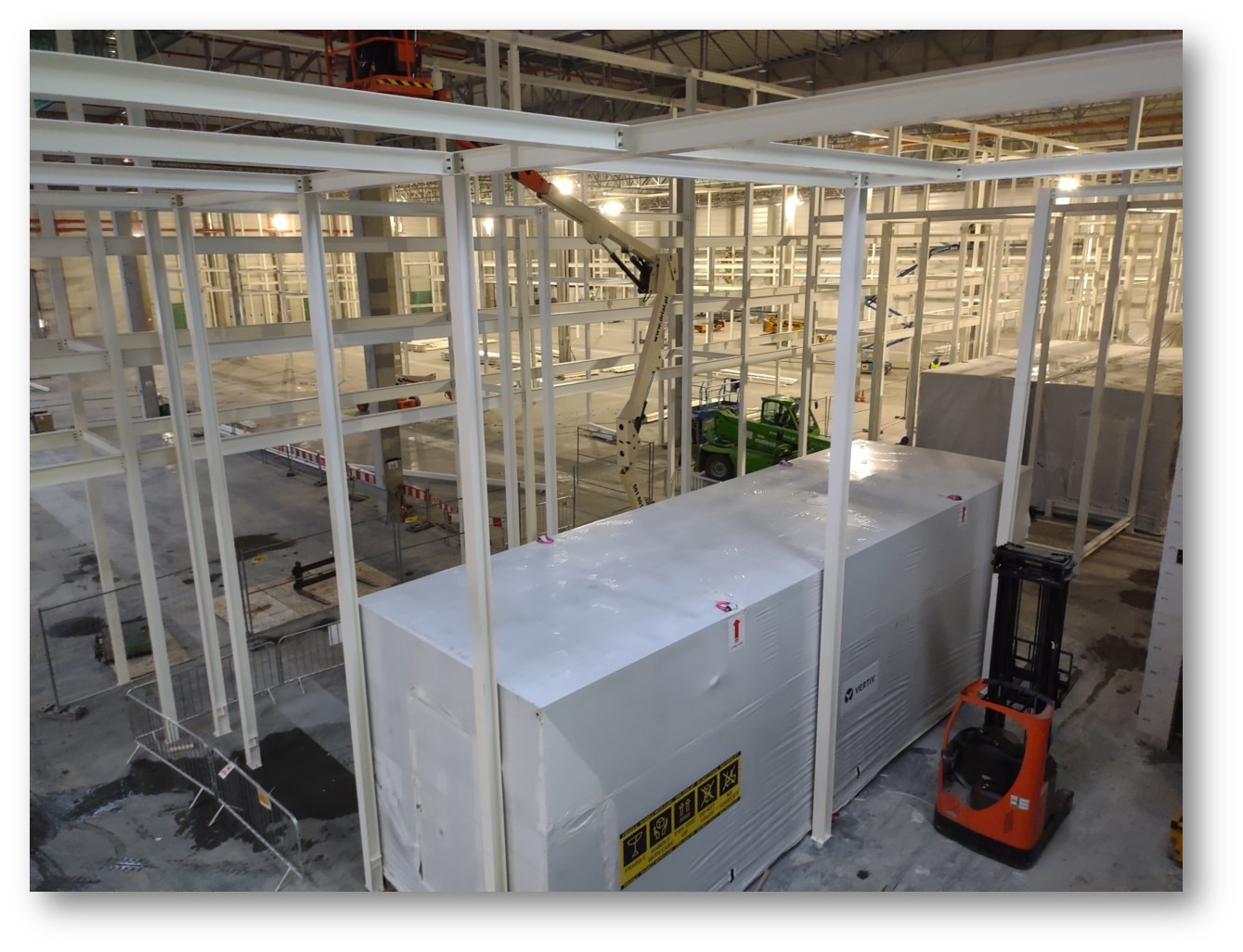 Wnętrze hali w trakcie budowy w Warszawie, widoczne białe stalowe ramy konstrukcyjne, zapakowany moduł i wózek widłowy, a w tle podnośniki koszowe.