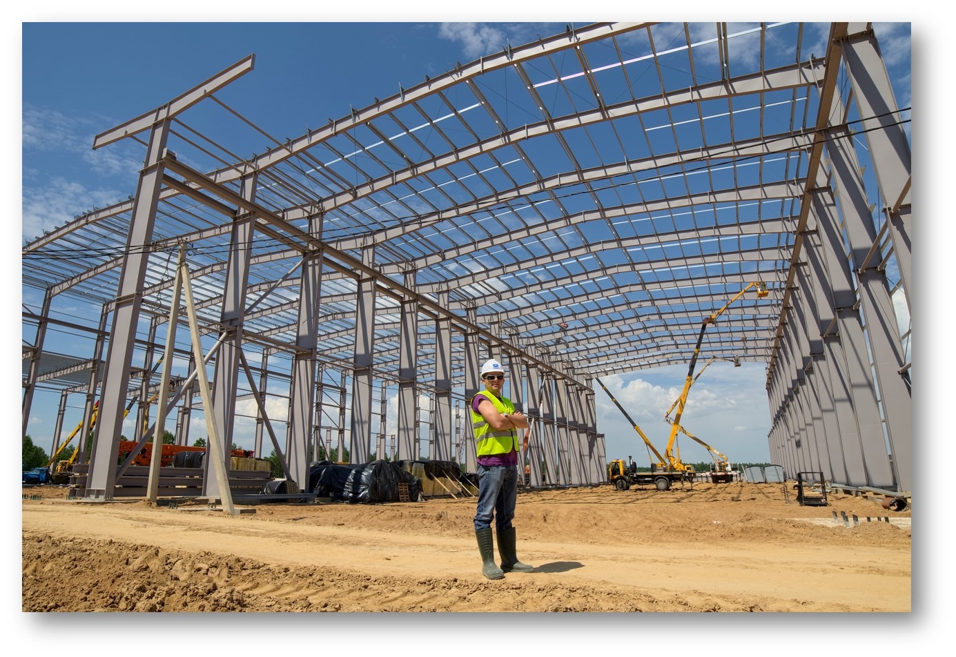 Montaż szkieletu hali stalowej z widocznym pracownikiem w kasku i kamizelce odblaskowej na tle nieba i maszyn budowlanych.