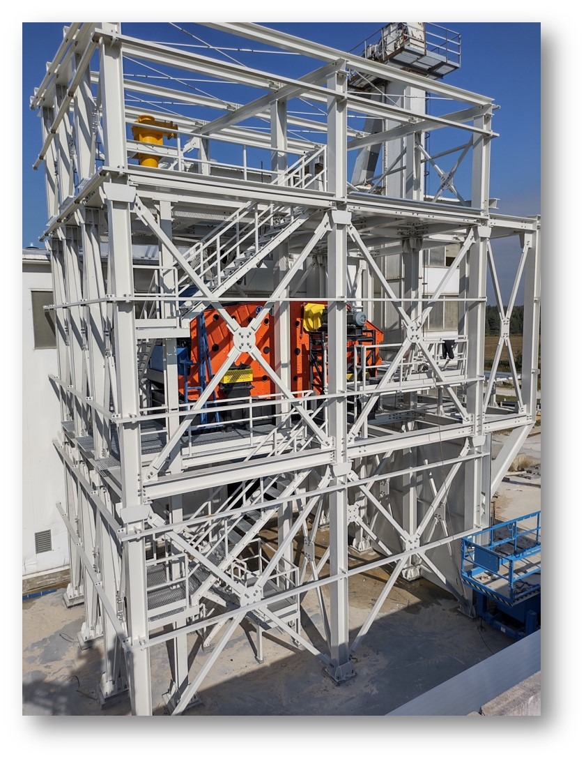 Wielopoziomowa konstrukcja stalowa w kolorze białym z pomarańczowym urządzeniem wewnątrz, widoczne schody i elementy złączne, na zewnątrz budynku w słoneczny dzień.