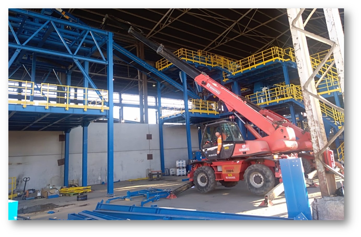 Montaż niebieskiej stalowej konstrukcji hali z żółtymi barierkami, widoczny czerwony wózek widłowy z operatorem w pomarańczowej kamizelce.