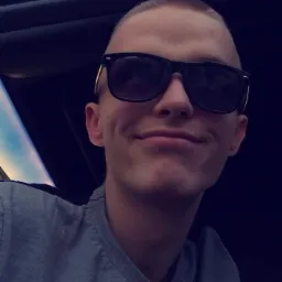 Portret uśmiechniętego młodego mężczyzny w okularach przeciwsłonecznych, z krótkimi włosami, siedzącego w samochodzie.