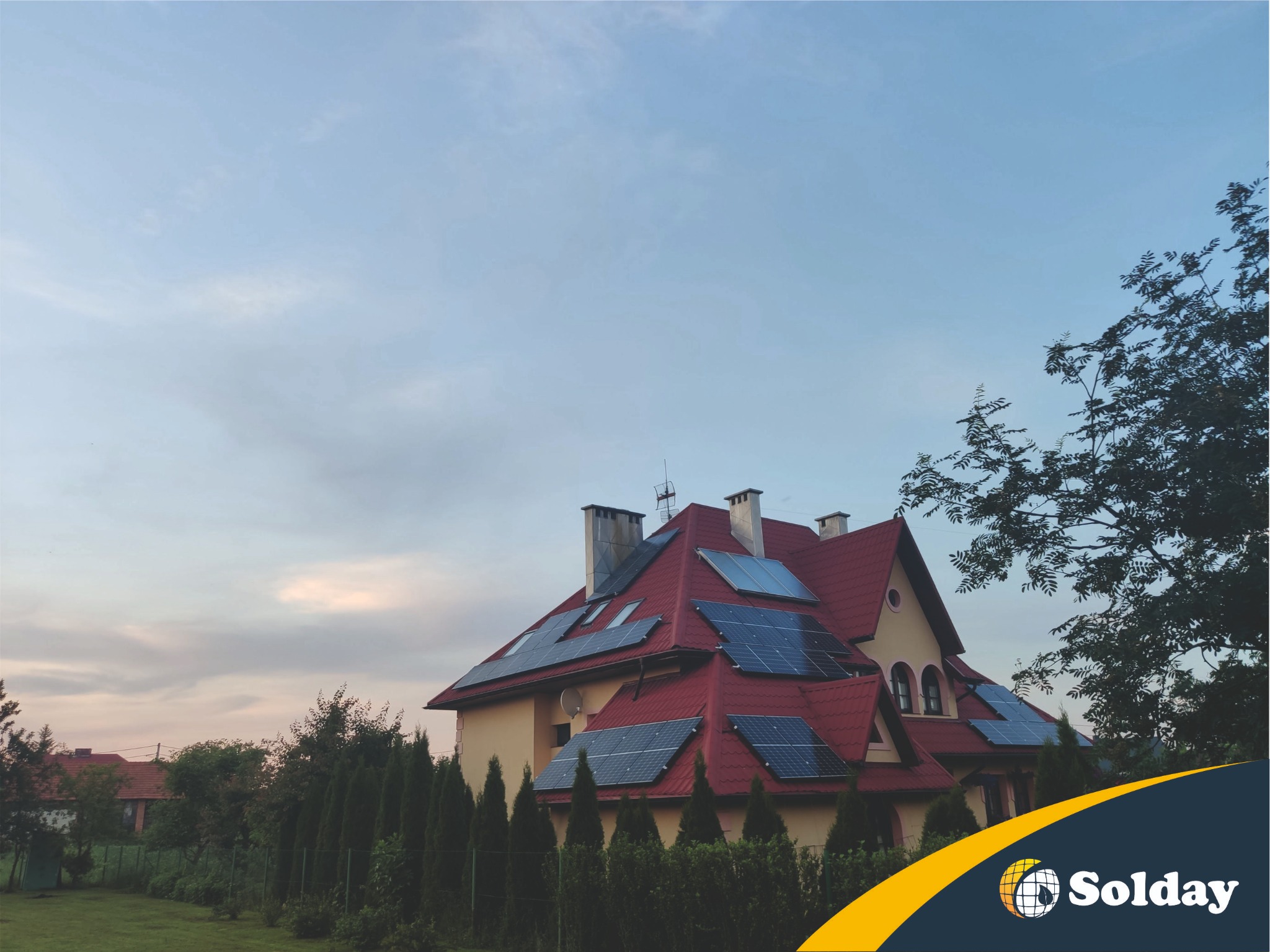 Dom jednorodzinny z panelami słonecznymi na czerwonym dachu, widok z ogrodu w pochmurny dzień, logo firmy w prawym dolnym rogu.