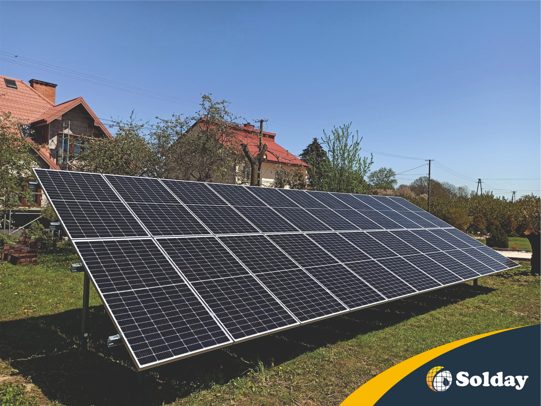 Panel słoneczny na trawniku przed domem z czerwoną dachówką w słoneczny dzień.