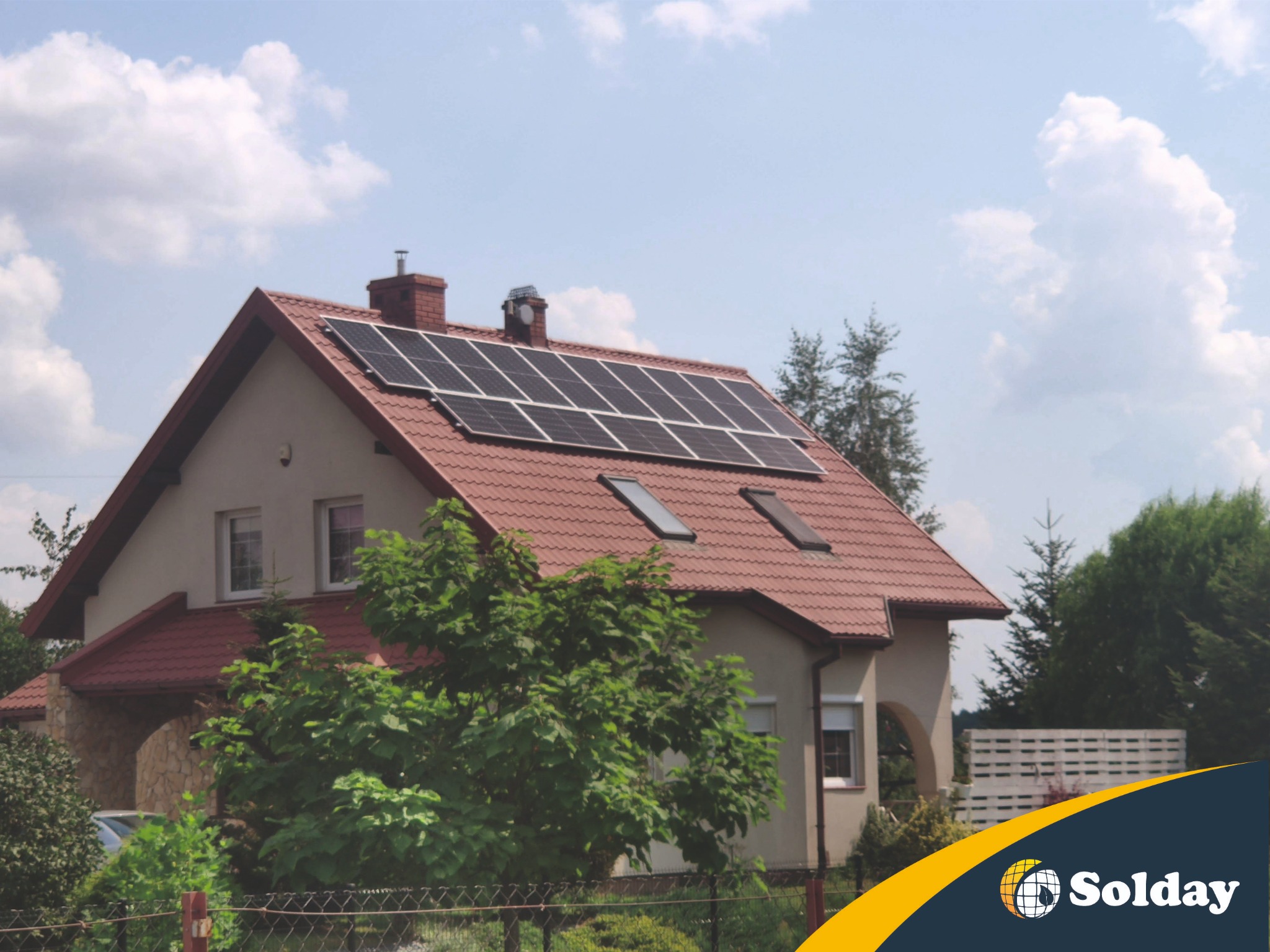 Dom jednorodzinny z panelami słonecznymi na czerwonym dachu i dwoma oknami dachowymi, widok z poziomu ogrodu, logo firmy w prawym dolnym rogu.