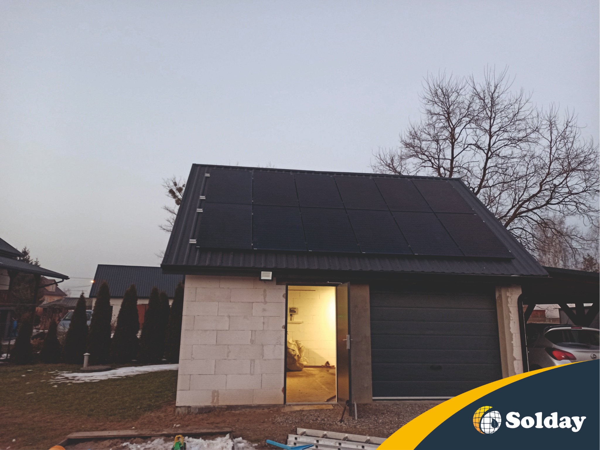 Dom z garażem, na dachu którego zamontowano panele fotowoltaiczne; widoczne otwarte drzwi garażu i drabina oparta o ścianę.