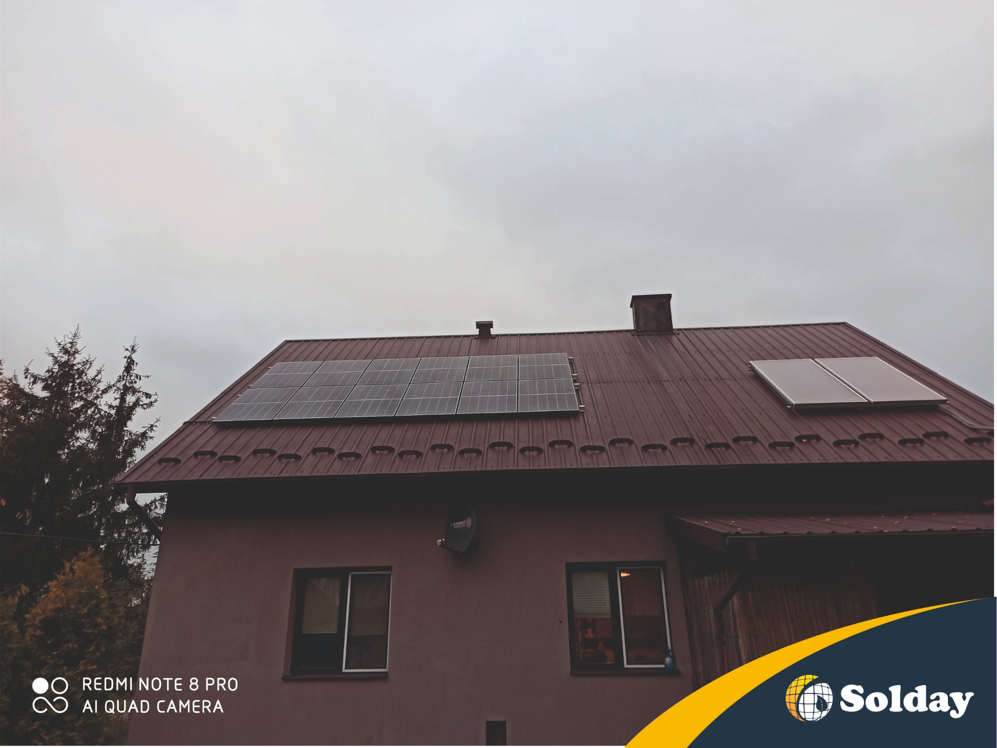 Dom jednorodzinny z panelami słonecznymi na dachu z blachy trapezowej w pochmurny dzień. Widoczna antena satelitarna na elewacji budynku.