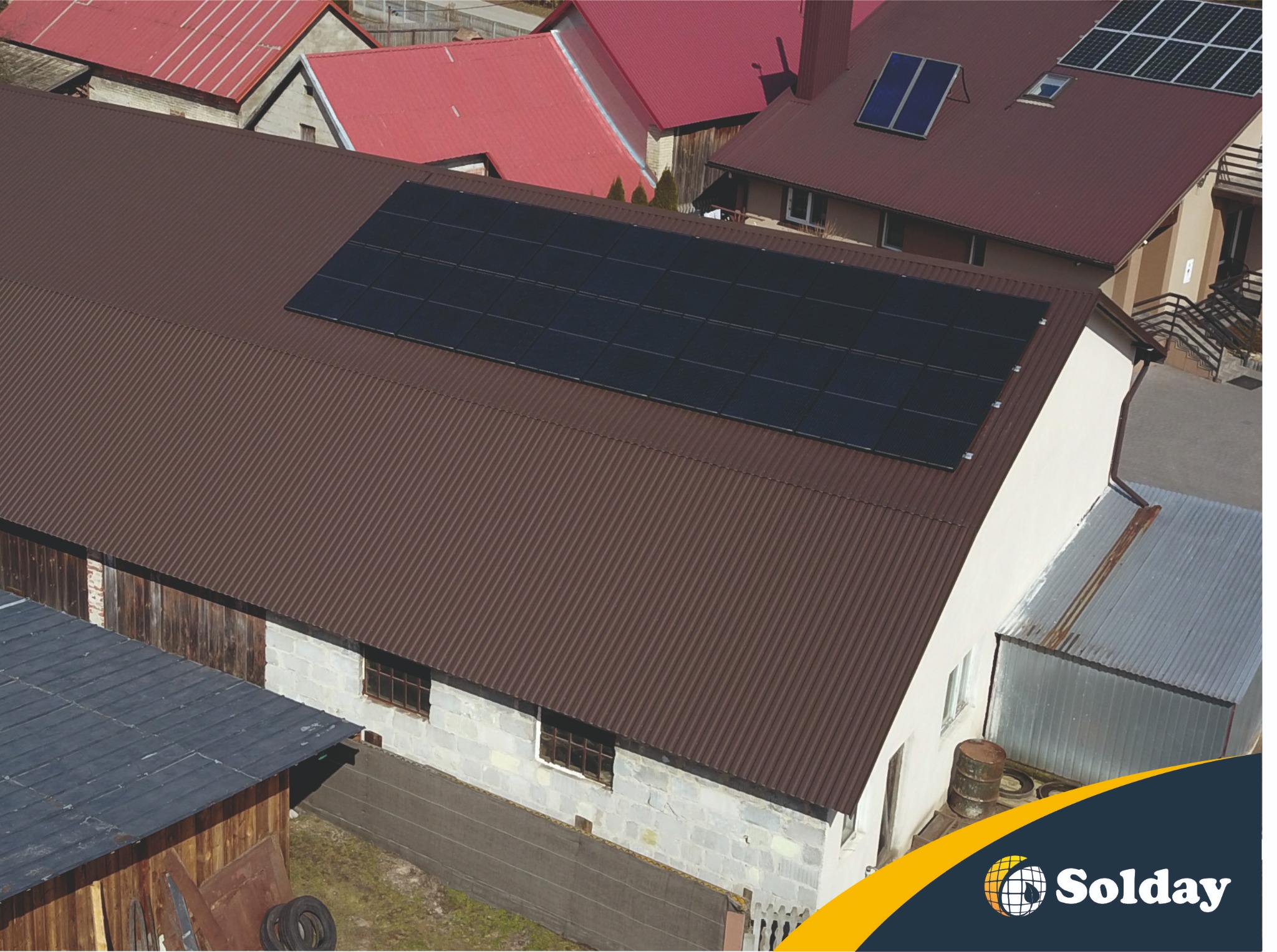 Widok z góry na instalację paneli słonecznych na brązowym, blaszanym dachu budynku gospodarczego, w tle inne budynki z czerwonymi dachami i logo firmy w prawym dolnym rogu.