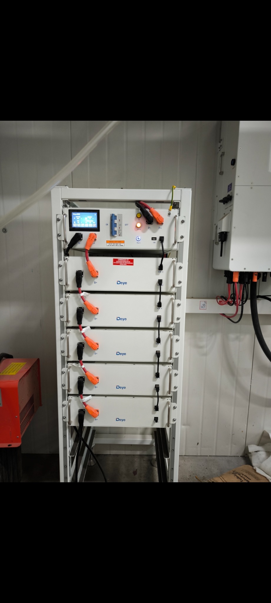 Biała szafa z modułami bateryjnymi Deye, podłączona do falownika, w pomieszczeniu technicznym z blach falistych.