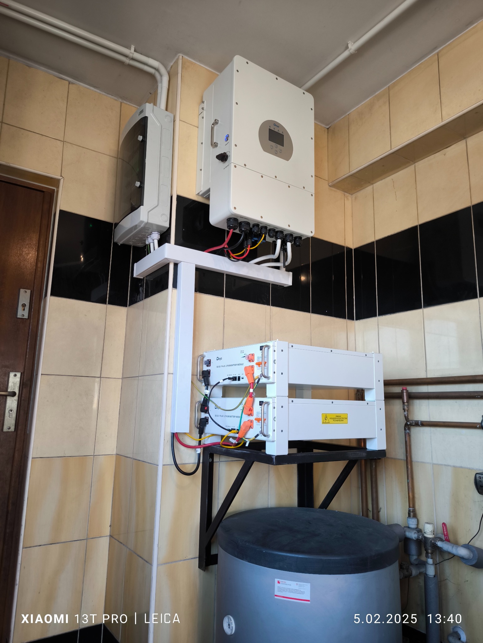 Dwie białe baterie litowo-żelazowo-fosforanowe marki Deye zamontowane na metalowym stelażu nad szarym zbiornikiem, podłączone do białego falownika na ścianie wyłożonej płytkami ceramicznymi...