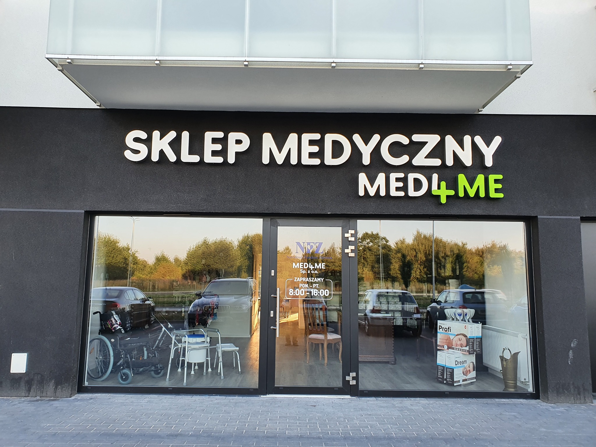 Fasada sklepu medycznego MED4ME z widocznym asortymentem: wózki inwalidzkie, krzesła toaletowe oraz pudełka z artykułami higienicznymi, odbijające się w szybach zaparkowane samochody.