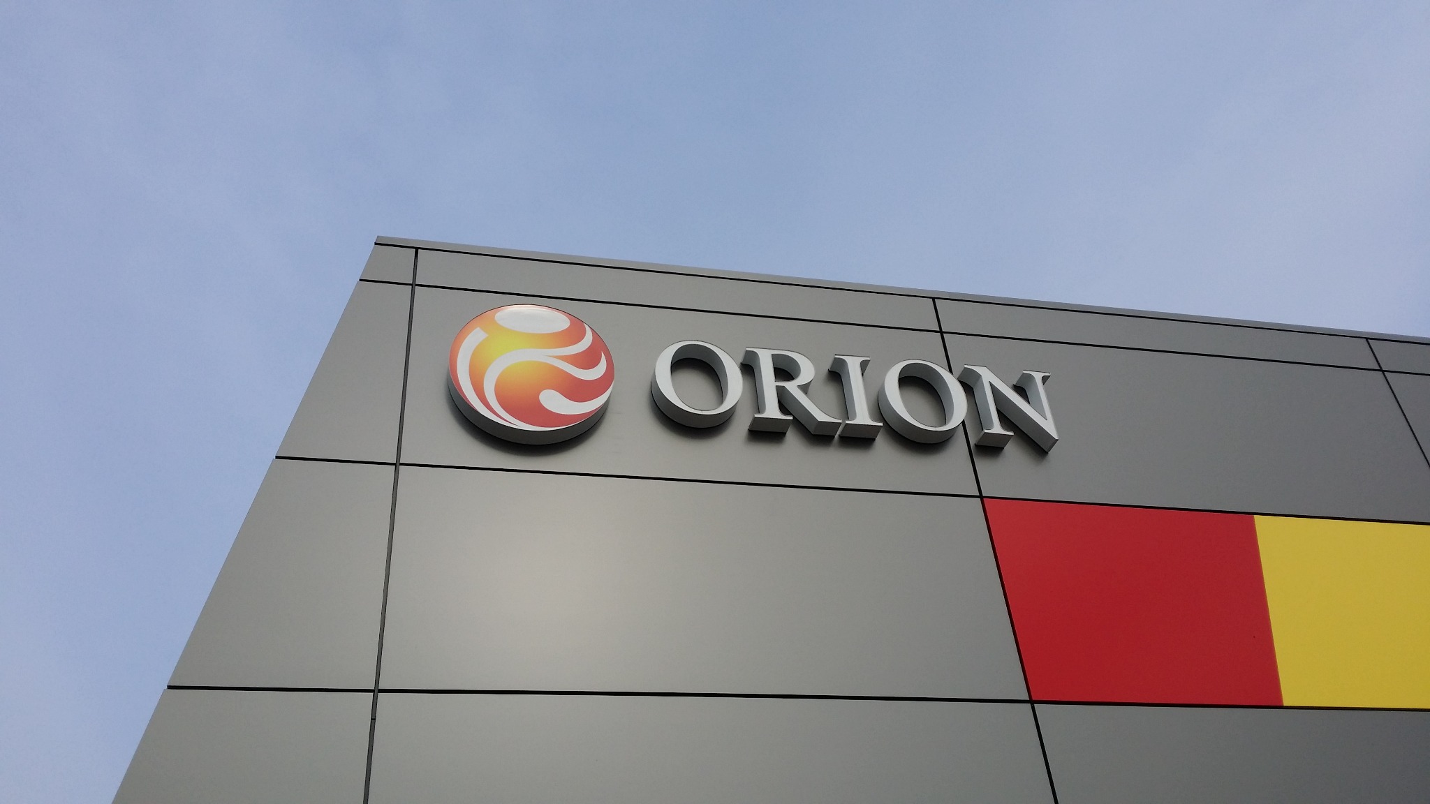 Elewacja budynku z logo i nazwą firmy 'ORION' w kolorze srebrnym, z dodatkowymi panelami w kolorach czerwonym i żółtym, na tle błękitnego nieba.