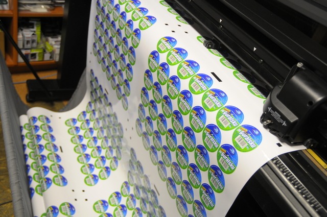 Arkusz z powtarzalnymi, okrągłymi etykietami z logo 'Góralsku' w kolorach zieleni i błękitu, częściowo pocięty przez ploter tnący, widoczne elementy maszyny drukującej.