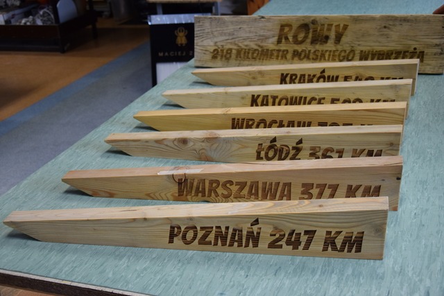 Drewniane drogowskazy z wypalonymi nazwami miast i odległościami w kilometrach, ułożone rzędami na stole, w tle tablica z napisem 'Rowy 218 kilometr polskiego wybrzeża'.