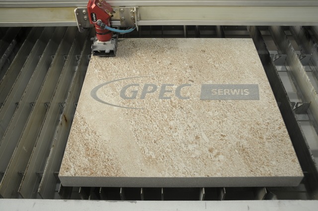 Proces cięcia wodą płytki ceramicznej z wygrawerowanym logo firmy GPEC Serwis za pomocą precyzyjnej maszyny. Widoczna głowica tnąca i stalowa kratownica pod płytką.