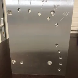 Metalowa płyta z nawierconymi otworami o różnych średnicach i oznaczeniami kątowymi od 0 do 40 stopni, leżąca na ciemnym blacie.