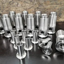 Srebrne, metalowe elementy z gwintem i otworami po obróbce skrawaniem, ułożone na ciemnym blacie na tle płyty drewnopodobnej.