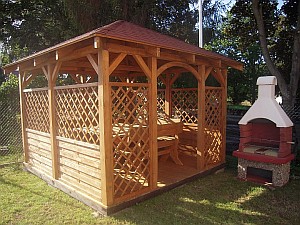 Drewniana altana ogrodowa z czerwonym dachem, ażurowe ścianki, stół i ławki wewnątrz, obok murowany grill z kominem.