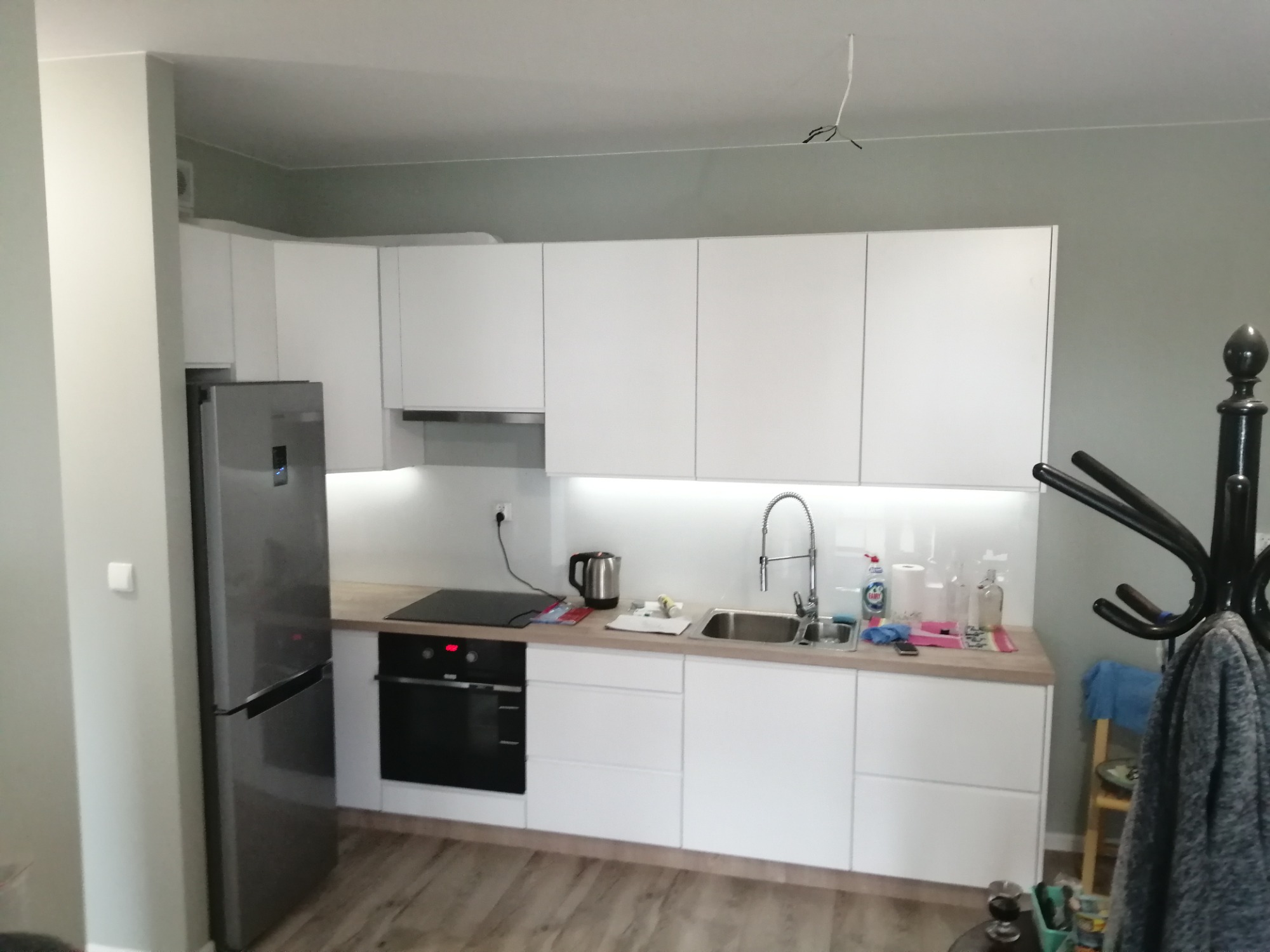 Jasna kuchnia z białymi frontami, srebrną lodówką i drewnianym blatem, widok na strefę gotowania i zlew, z wiszącymi kablami na suficie i wieszakiem na ubrania w rogu.