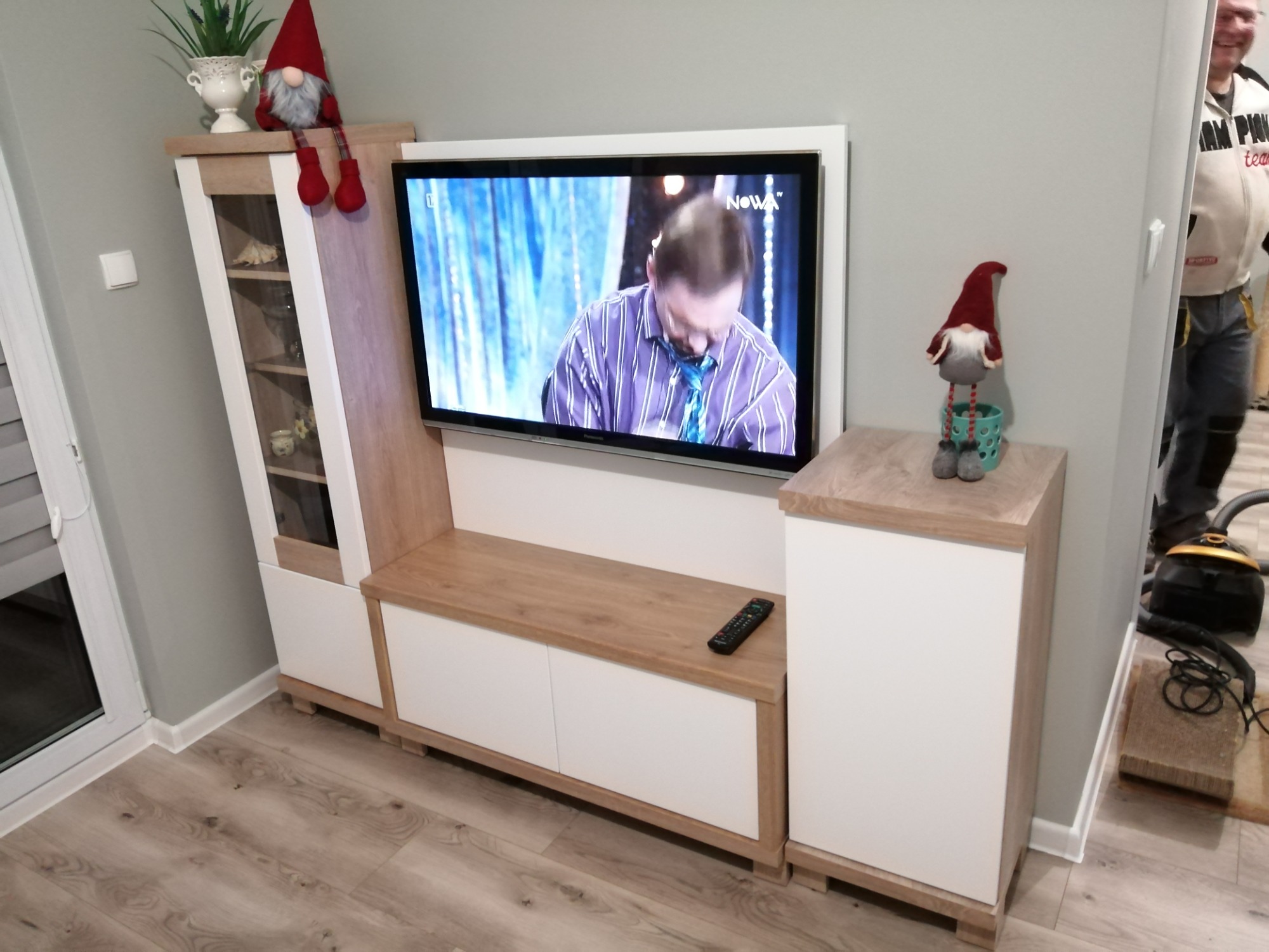 Zestaw mebli RTV w odcieniach bieli i drewna z telewizorem, ozdobiony figurkami skrzatów, na podłodze widoczny odkurzacz.