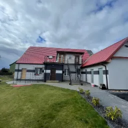 Dwukondygnacyjny budynek z czerwonym dachem z blachodachówki, szarą elewacją z poziomymi czarnymi pasami i zewnętrznymi schodami; widoczny trawnik i chodnik z kostki.