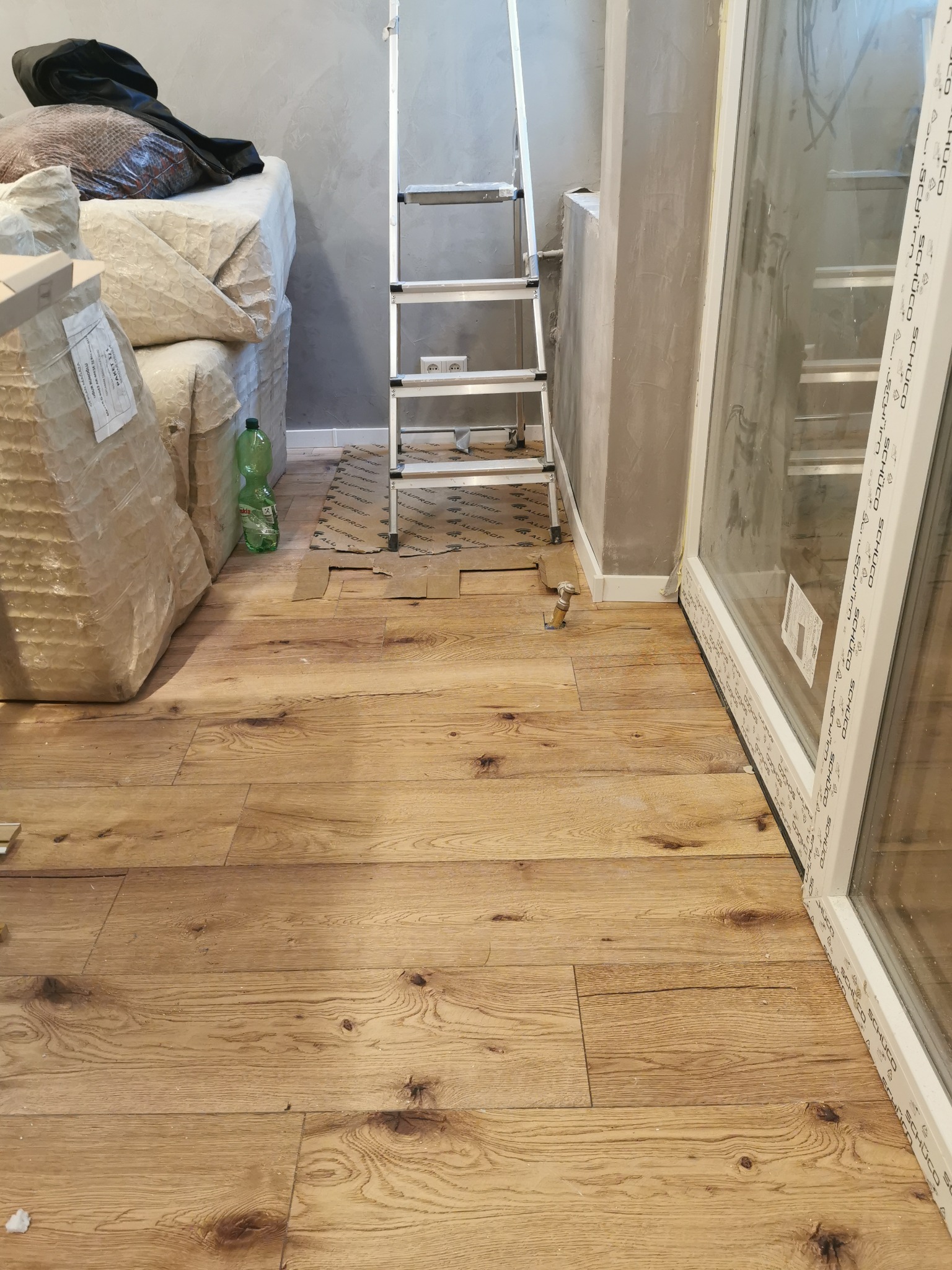 Podłoga z paneli drewnopodobnych w trakcie montażu, widoczna drabina, okno z białym profilem, folia ochronna na podłodze i butelka po napoju.
