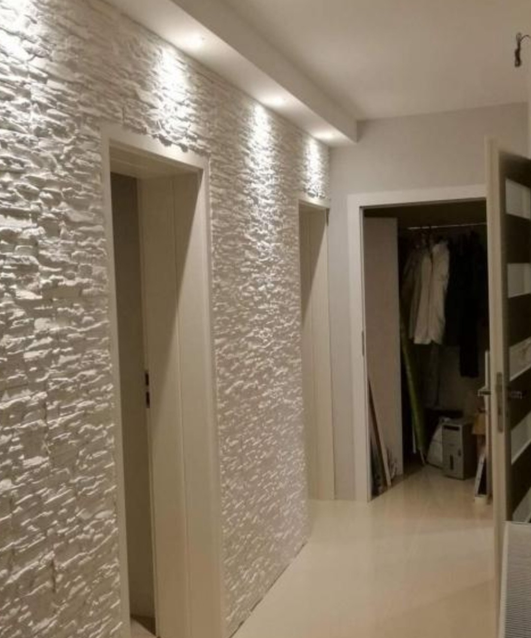 Korytarz z dekoracyjną, białą ścianą imitującą kamień, oświetloną punktowo, z widocznymi drzwiami i otwartą garderobą w tle.