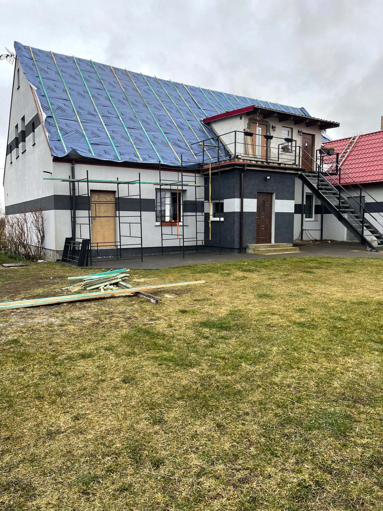 Dom w trakcie prac dekarskich, dach pokryty niebieską membraną i drewnianymi łatami, widoczne rusztowania i składowane deski na trawniku.