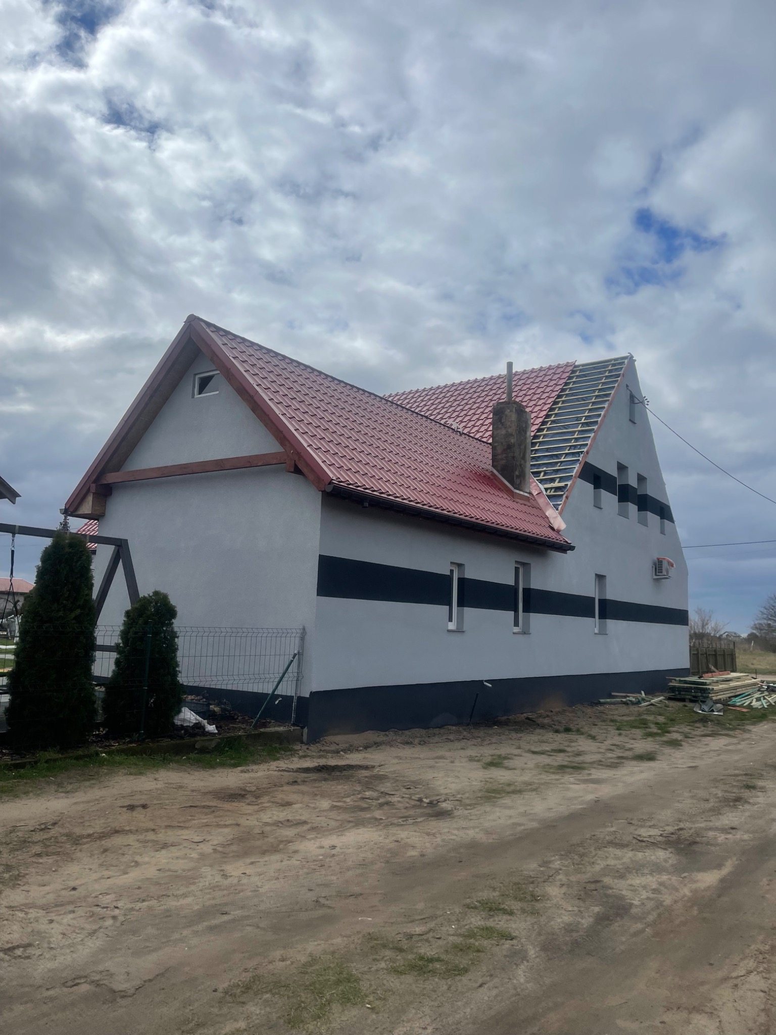 Dom jednorodzinny w trakcie remontu dachu, z częściowo ułożoną czerwoną dachówką i widoczną konstrukcją więźby dachowej. Elewacja w kolorze szarym z poziomymi czarnymi pasami. Widoczne okna, komin...