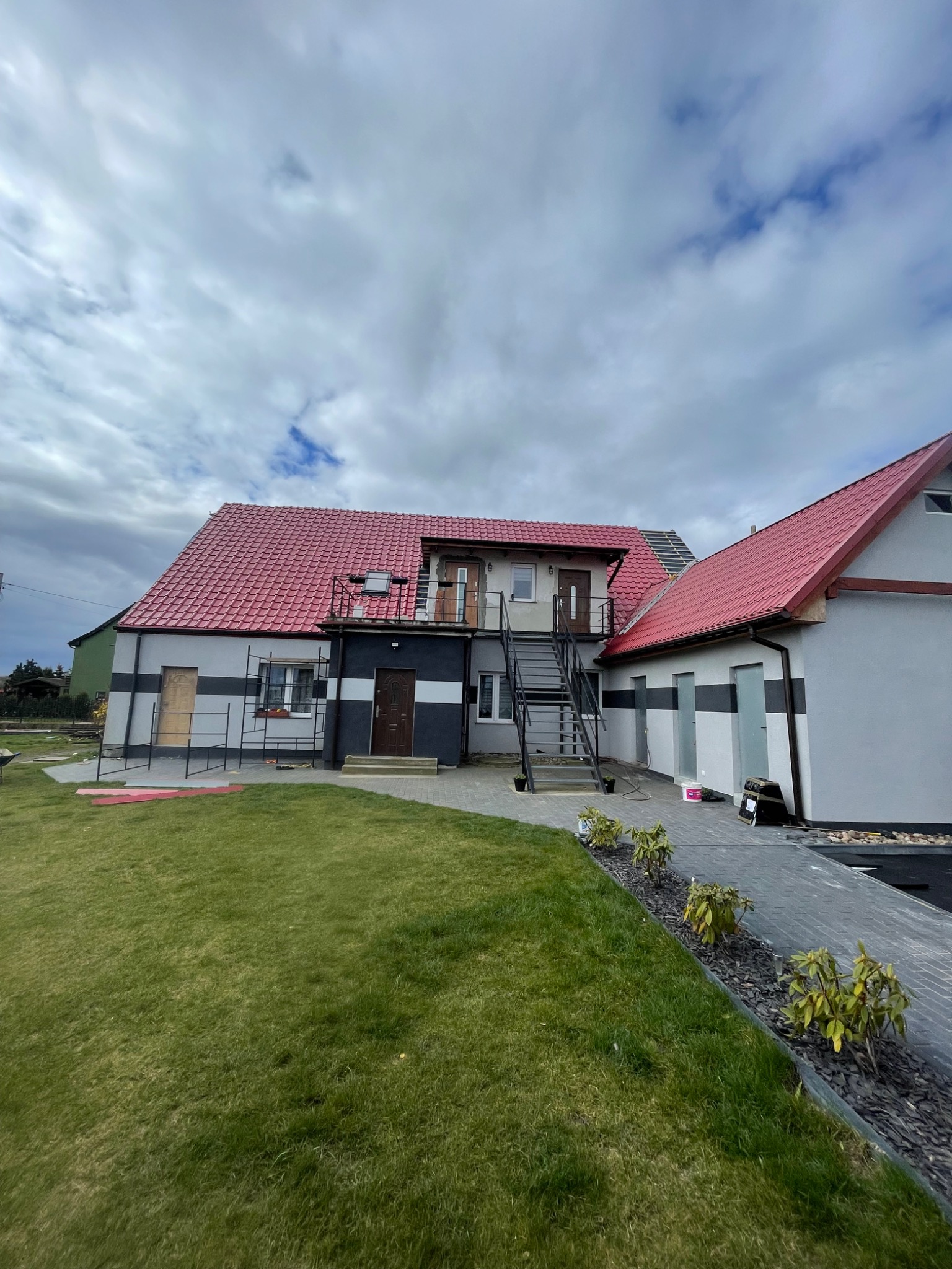 Dwukondygnacyjny budynek z czerwonym dachem z blachodachówki, szarą elewacją z poziomymi czarnymi pasami i zewnętrznymi schodami; widoczny trawnik i chodnik z kostki.