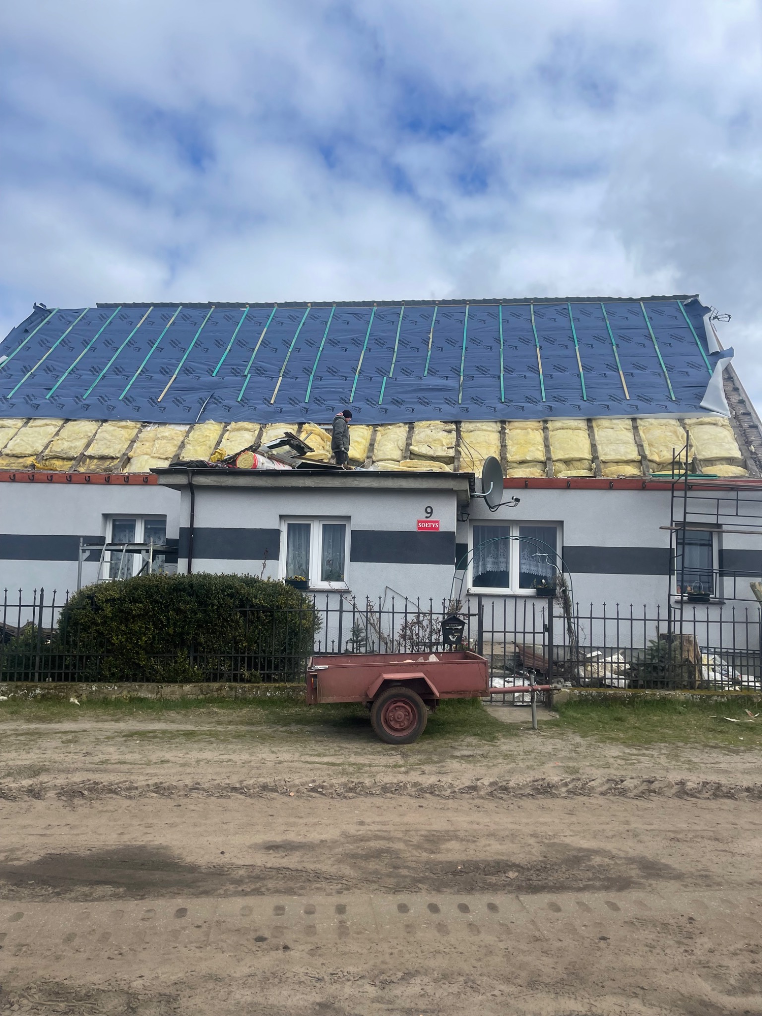 Remont dachu domu jednorodzinnego w trakcie prac: widoczna izolacja termiczna z wełny mineralnej, membrana dachowa i listwy kontrłat. Na dachu stoi pracownik w roboczym ubraniu. W tle budynek...