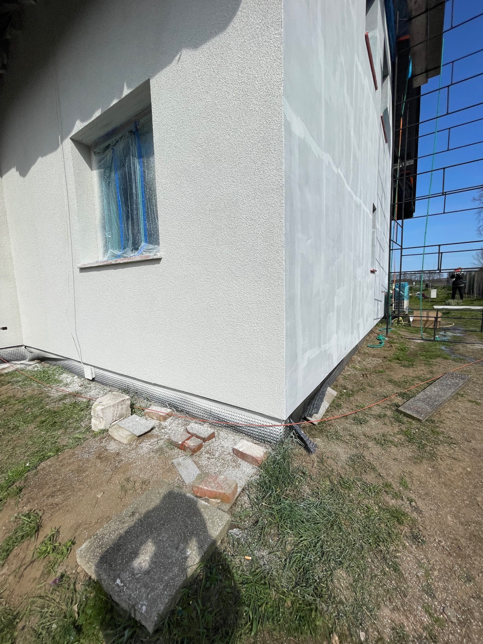 Fragment elewacji budynku po nałożeniu jasnego tynku strukturalnego, okno zabezpieczone folią malarską, widoczne rusztowanie i teren wokół budynku w trakcie prac wykończeniowych.