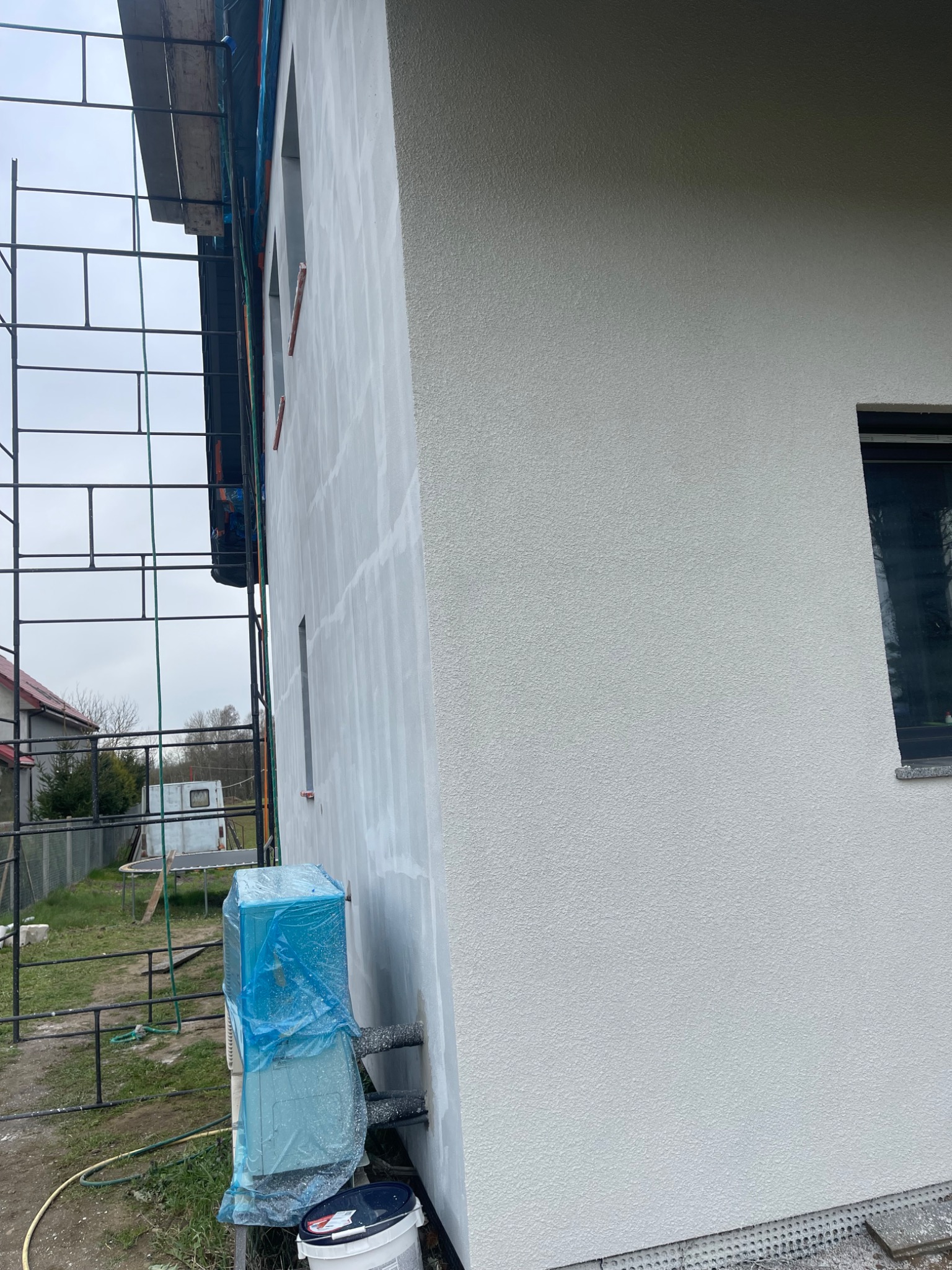 Świeżo otynkowana elewacja domu jednorodzinnego z widocznym rusztowaniem, zabezpieczonym agregatem klimatyzacji folią oraz pojemnikiem z farbą elewacyjną u dołu ściany.