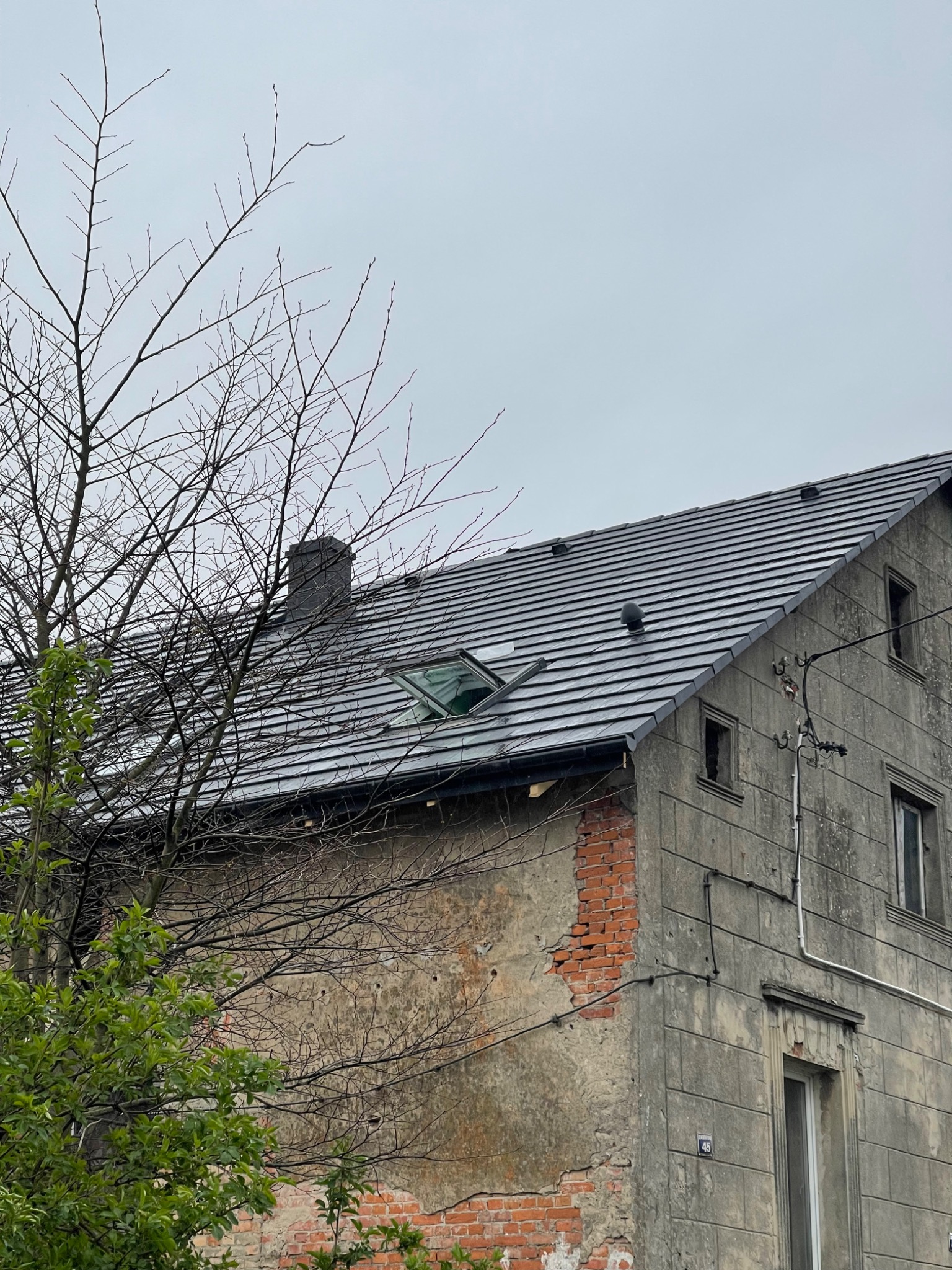 Dwupiętrowy budynek z szarą elewacją i nowym, ciemnoszarym dachem z otwartym oknem dachowym, widoczne fragmenty odsłoniętej cegły na elewacji, na pierwszym planie gałęzie drzewa.