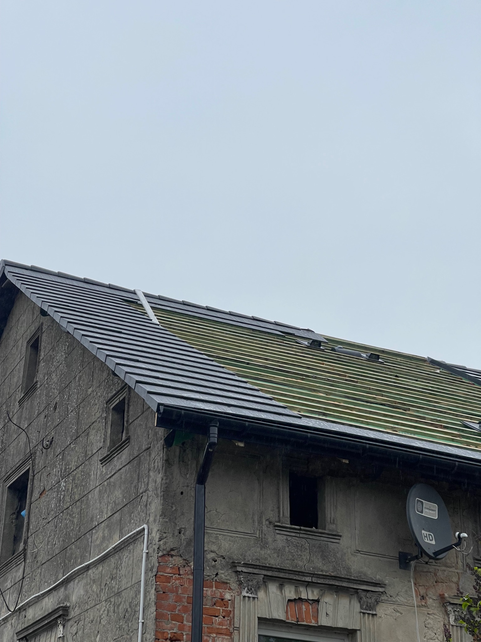 Częściowy widok dachu z czarną dachówką w trakcie układania na drewnianej konstrukcji, widoczny fragment starego, szarego budynku z anteną satelitarną.