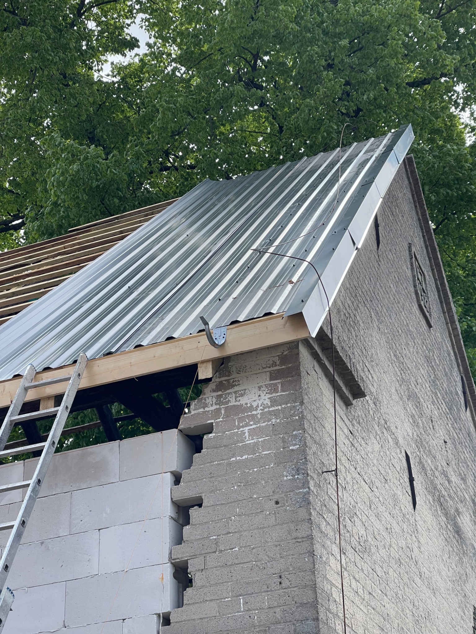 Montaż blachy trapezowej na dachu budynku z cegły, widoczne krokwie, łata, drabina oparta o ścianę. Częściowo otynkowana ściana z białych bloczków.