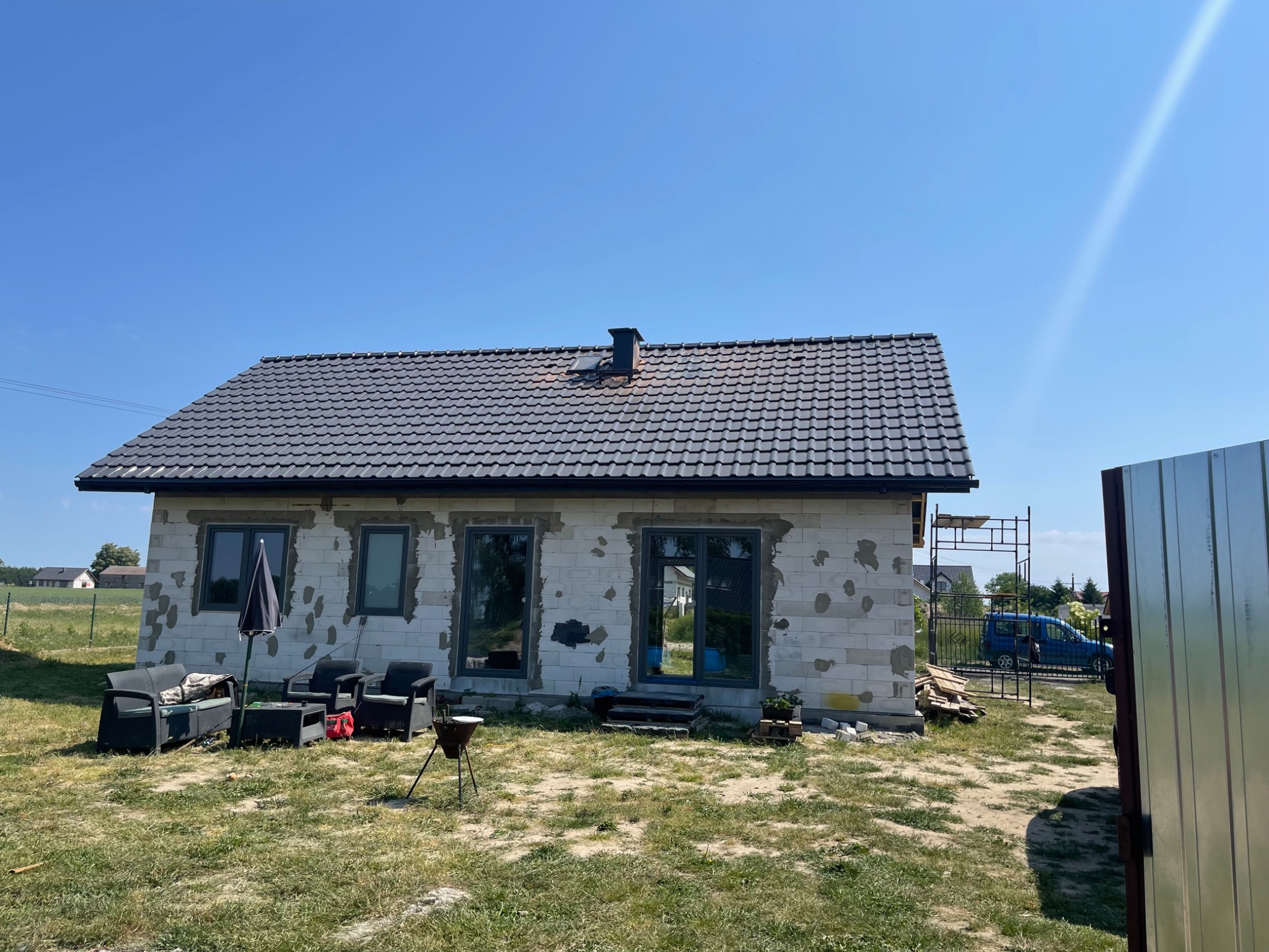 Dom jednorodzinny w trakcie budowy, z widocznymi fragmentami nieotynkowanych ścian z betonu komórkowego, ciemnoszara dachówka ceramiczna, okna i drzwi w kolorze grafitowym, na trawniku meble...