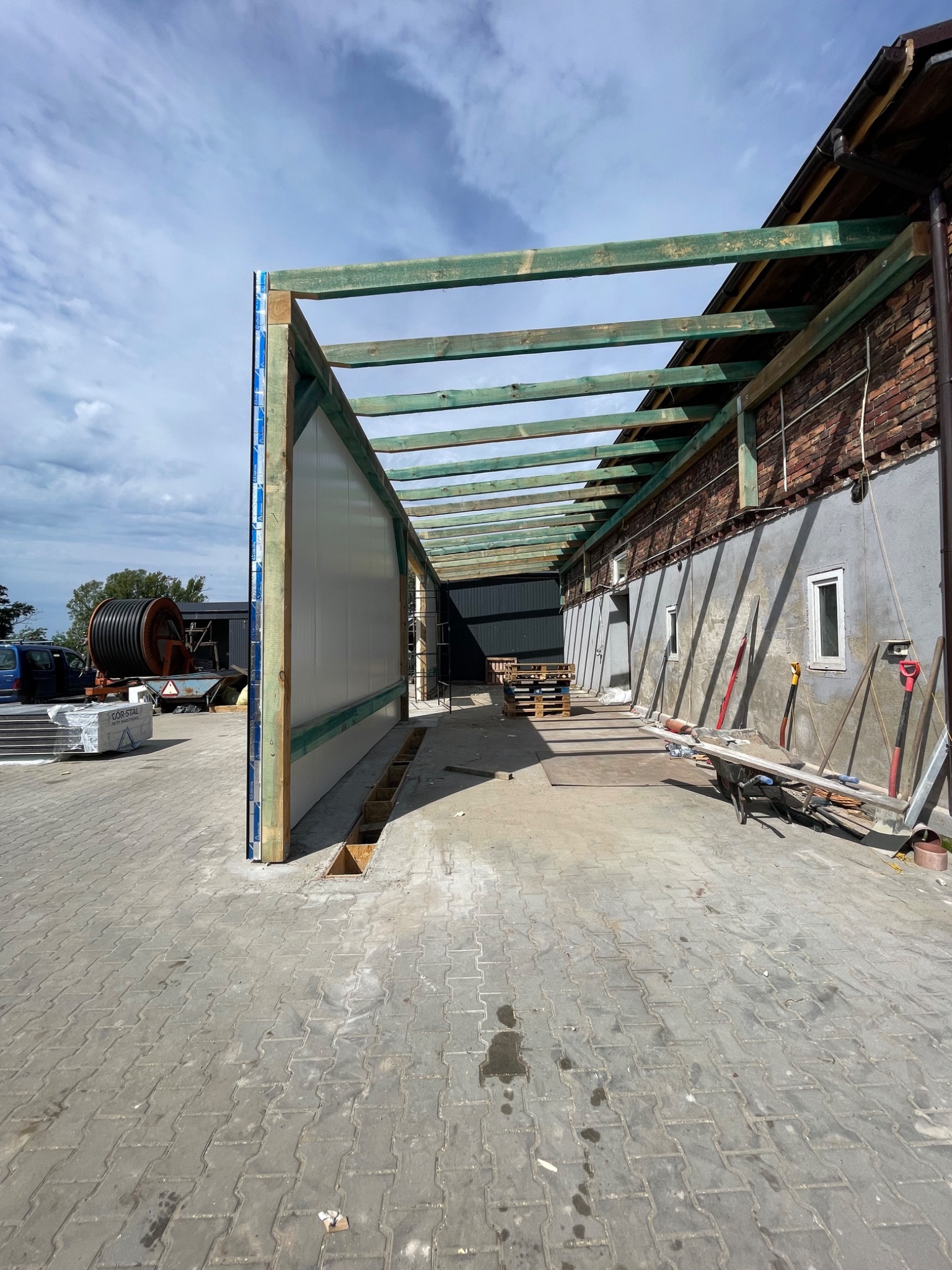 Budowa zadaszenia przy budynku z ceglaną ścianą, widoczne drewniane elementy konstrukcyjne i białe panele ścienne, narzędzia budowlane w tle.