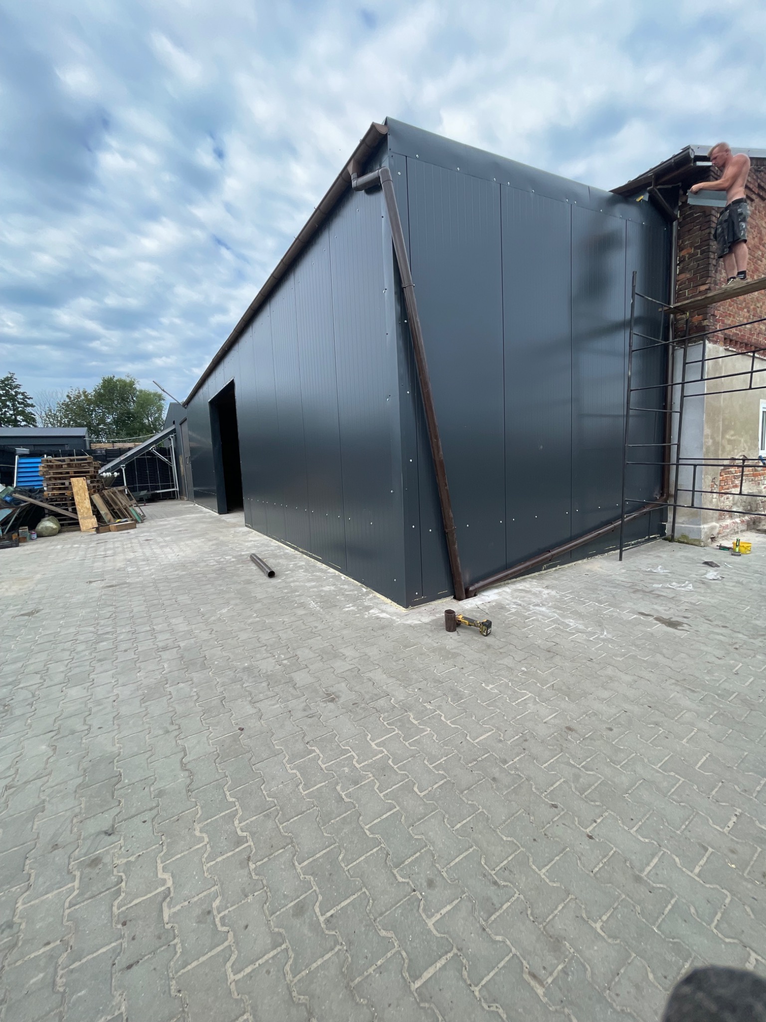 Montaż paneli elewacyjnych na budynku gospodarczym z blachy trapezowej, widoczny pracownik na rusztowaniu przy dachu oraz narzędzia na ziemi.