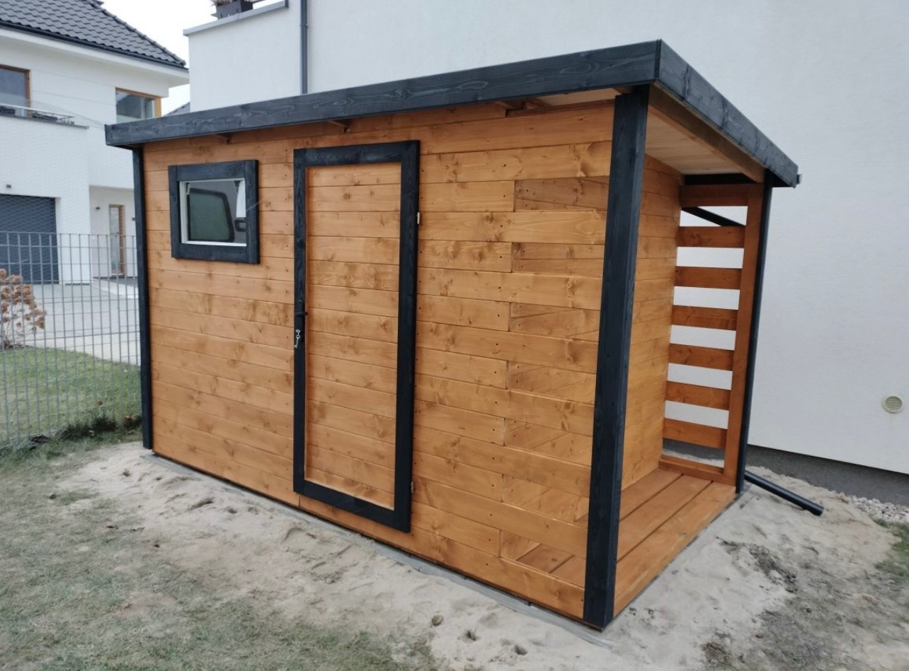 Drewniana szopa narzędziowa z czarnymi obramowaniami okna i drzwi, z widocznym fragmentem ogrodu i domu w tle.