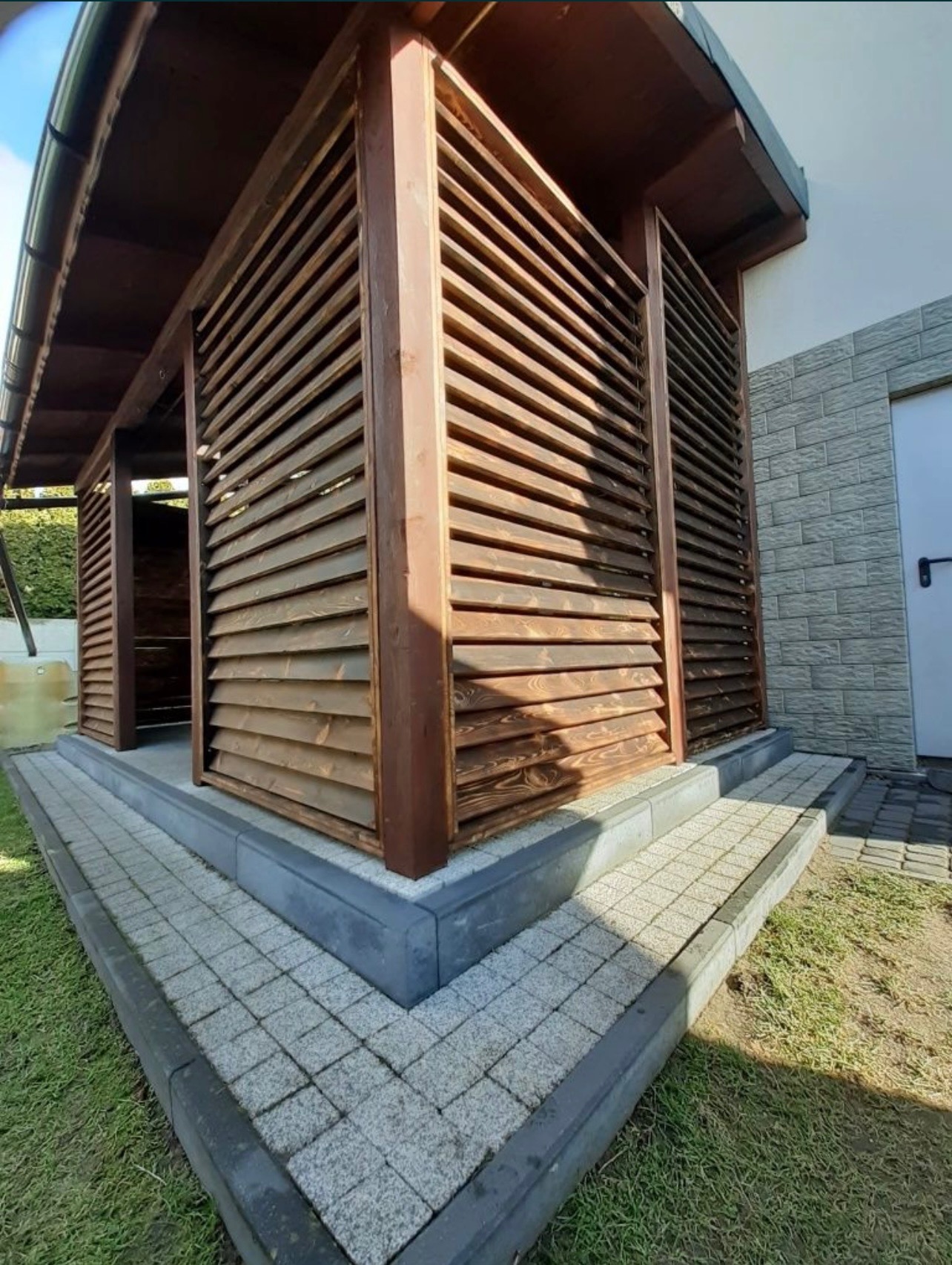 Drewniana konstrukcja z żaluzjami, prawdopodobnie altana lub zadaszenie tarasu, z kostki brukowej ułożony chodnik i schody.