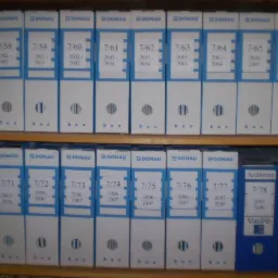 P&oacute;łka z dwoma rzędami segregator&oacute;w archiwizacyjnych marki Donau, ponumerowanych i oznaczonych rocznikami, z dwoma segregatorami VauPe w prawym dolnym rogu.