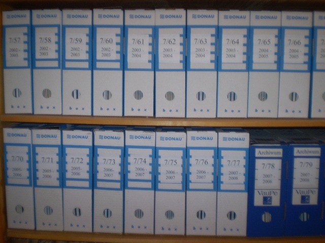Półka z dwoma rzędami segregatorów archiwizacyjnych marki Donau, ponumerowanych i oznaczonych rocznikami, z dwoma segregatorami VauPe w prawym dolnym rogu.