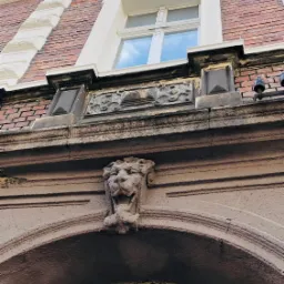 Fragment fasady kamienicy z dekoracyjnym lwem nad łukiem okiennym i zdobieniami pod oknem na tle ceglanej ściany.