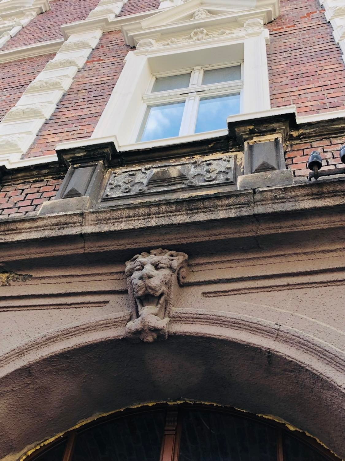 Fragment fasady kamienicy z dekoracyjnym lwem nad łukiem okiennym i zdobieniami pod oknem na tle ceglanej ściany.