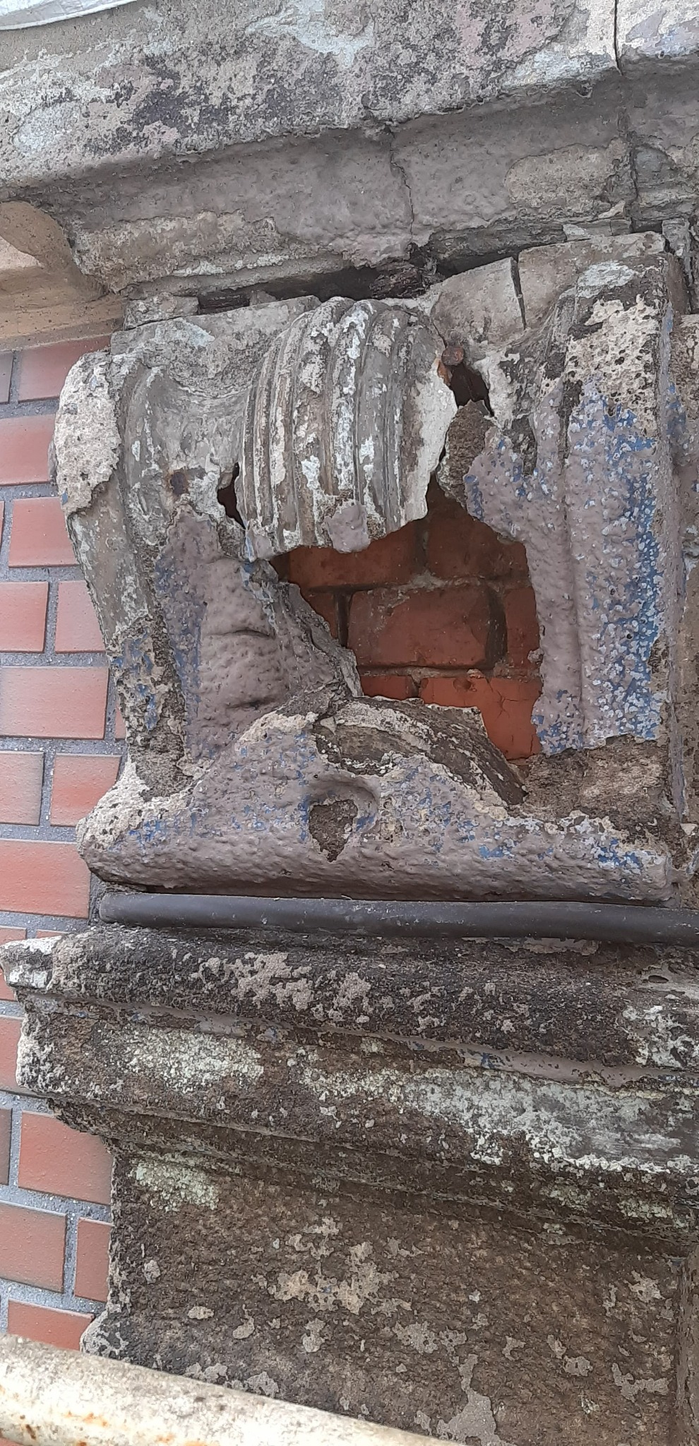 Zniszczony fragment dekoracyjnej elewacji z ubytkiem odsłaniającym czerwoną cegłę, z widocznymi śladami starej farby i erozji.