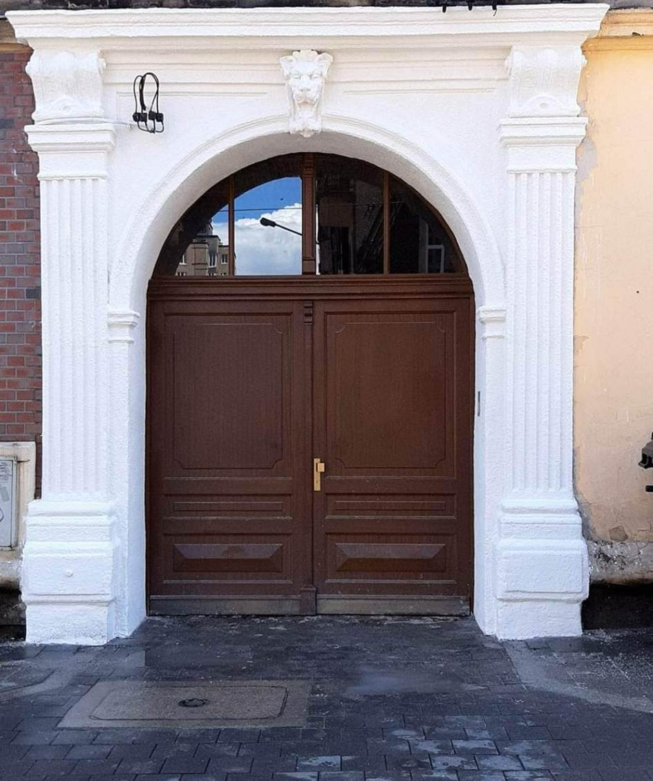 Odnowione wejście do budynku z drewnianymi drzwiami, łukiem i kolumnami pomalowanymi na biało, zwieńczone rzeźbą lwa.