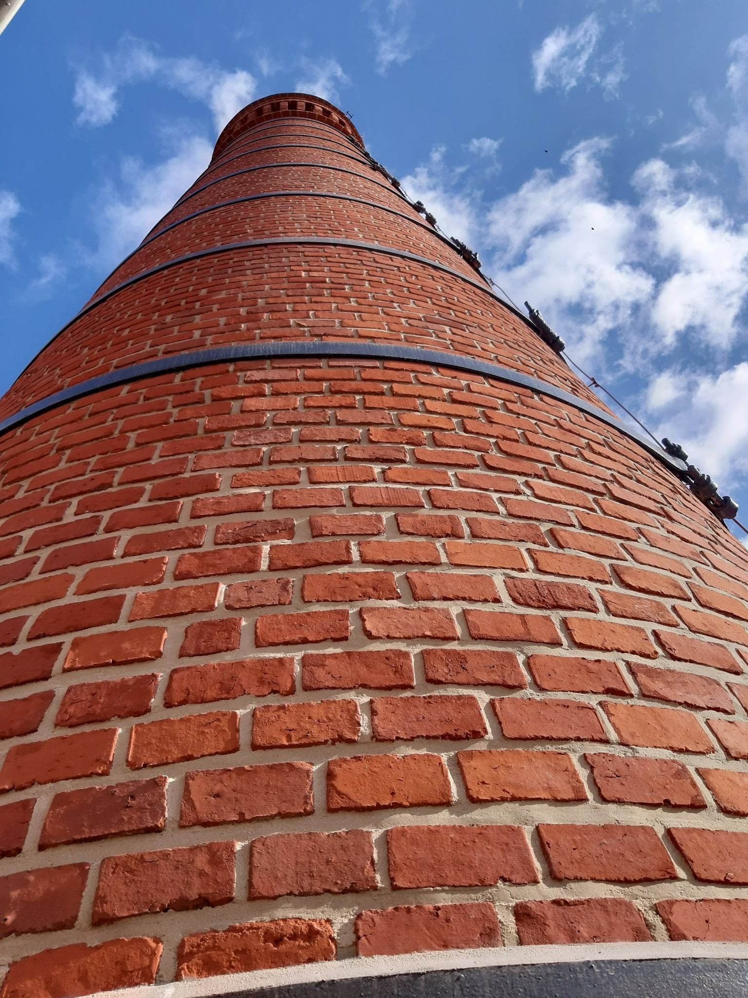 Wysoka, ceglana konstrukcja komina z metalowymi obręczami, widziana z dołu na tle błękitnego nieba z chmurami.