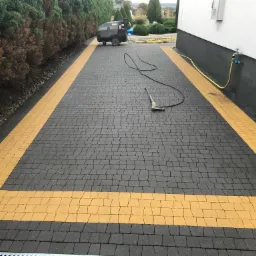Ciemnoszara kostka brukowa z żółtymi pasami obrysowującymi obszar, z widocznym urządzeniem wysokociśnieniowym Kärcher i lancą do mycia, w tle żywopłot i fragment budynku.