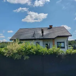 Pracownik w uprzęży na drabinie oparty o dach domu jednorodzinnego z ciemnej dachówki, wykonuje prace na wysokości na tle błękitnego nieba z chmurami.