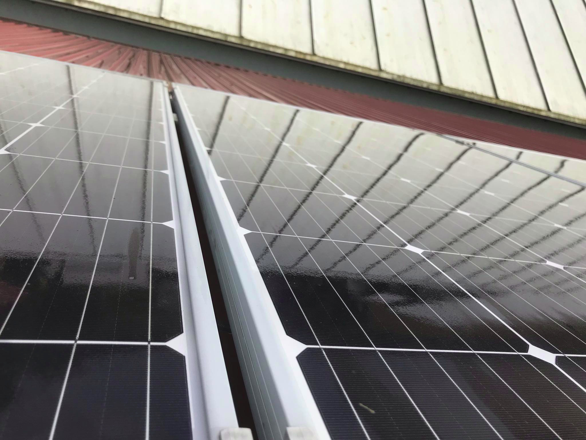 Błyszczące panele słoneczne zamontowane na czerwonym dachu z blachy trapezowej, widoczne białe ramy paneli i odbicie konstrukcji dachu w ich powierzchni.